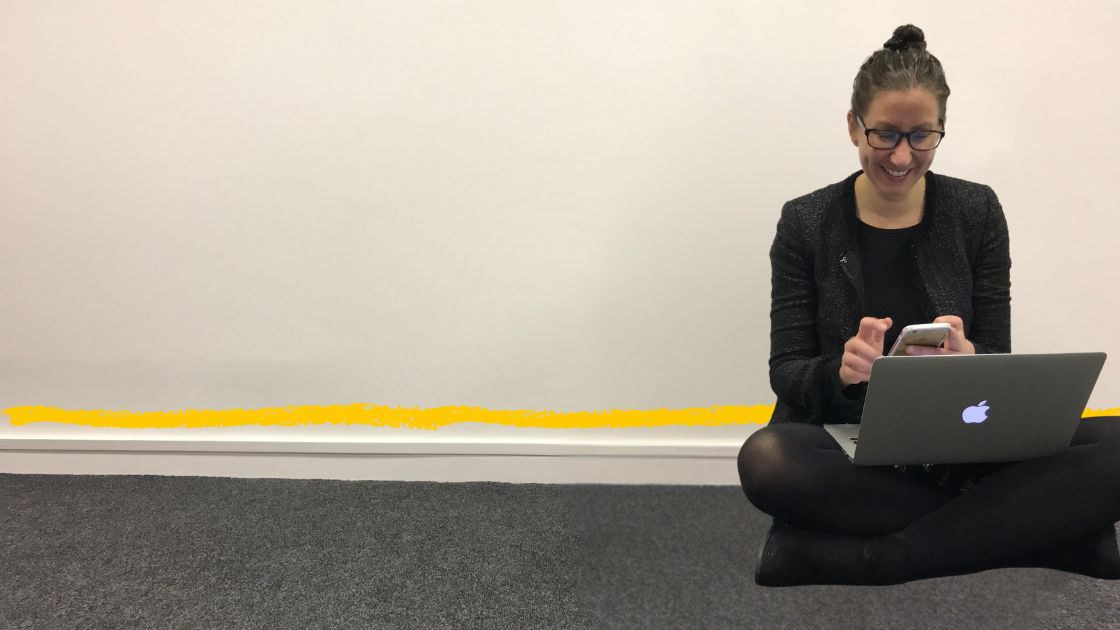 A woman with a MacBook on her knees, sitting cross-legged on the floor in an office