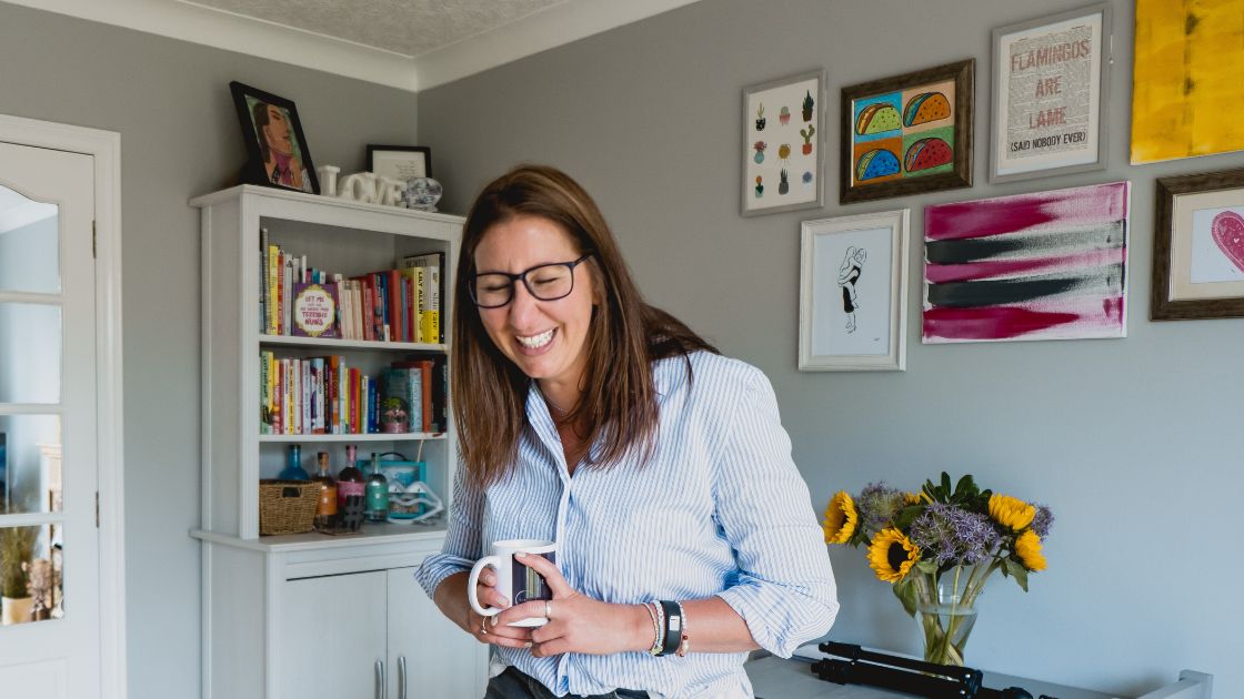 a photo of Suz Bird leaning against a table, holding a coffee mug and laughing